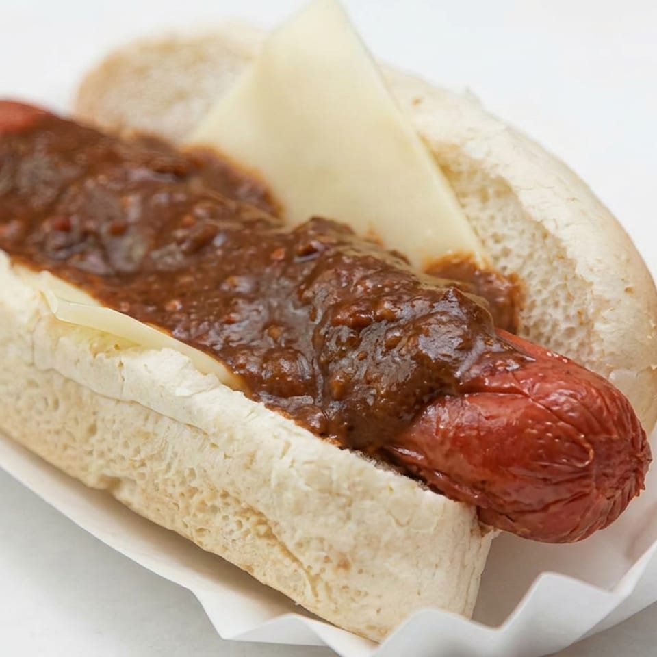 Chili cheese hot dog in a soft bun, topped with melted cheese and thick chili sauce on a paper tray.